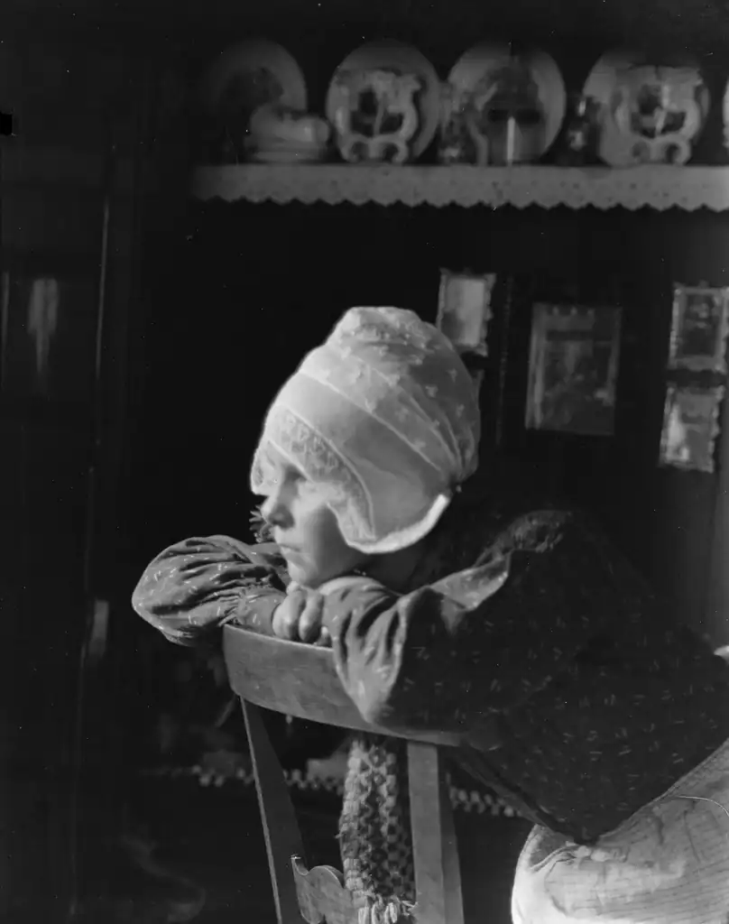 Child praying with head bowed, wearing a bonnet in a cozy home setting in the Netherlands