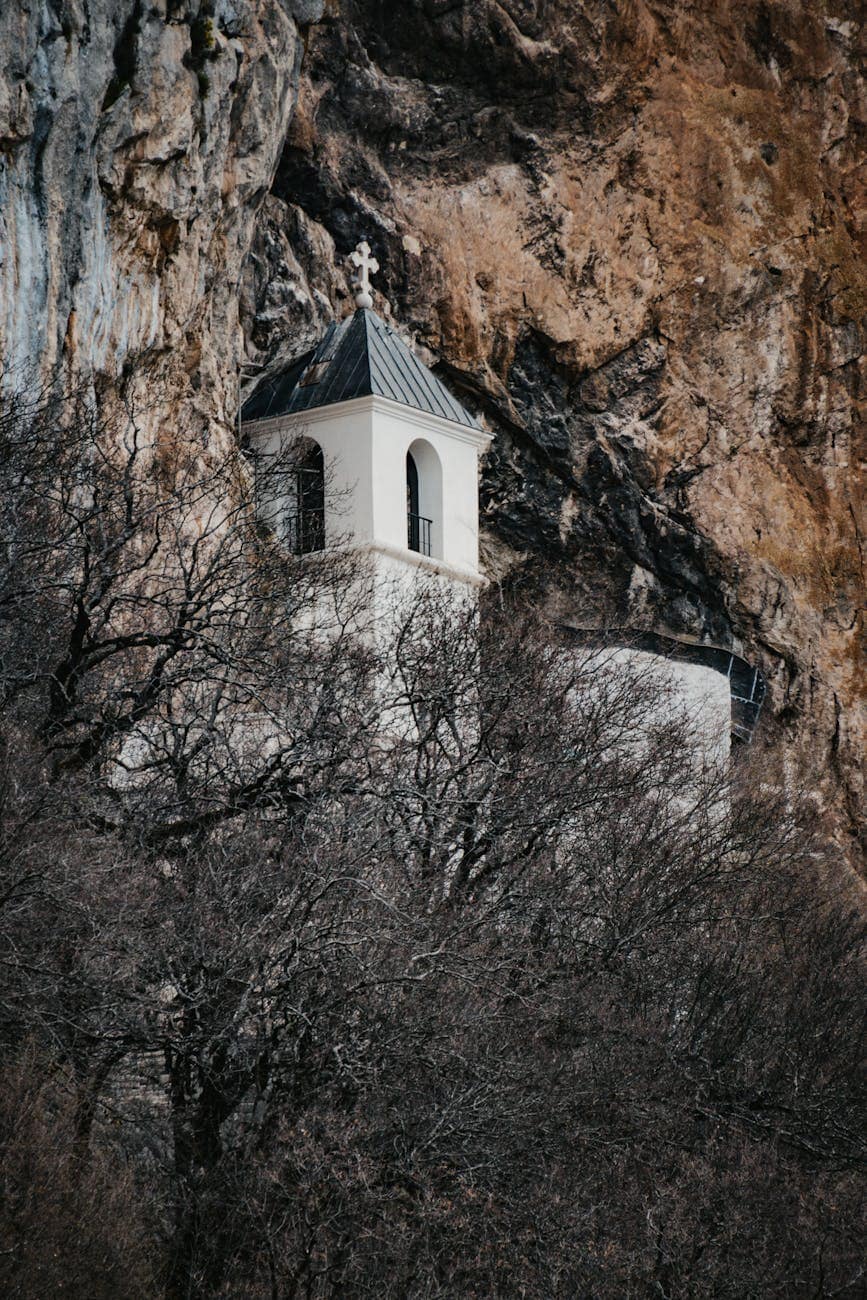 Small white chapel on rocky mountain cliff with trees in foreground.