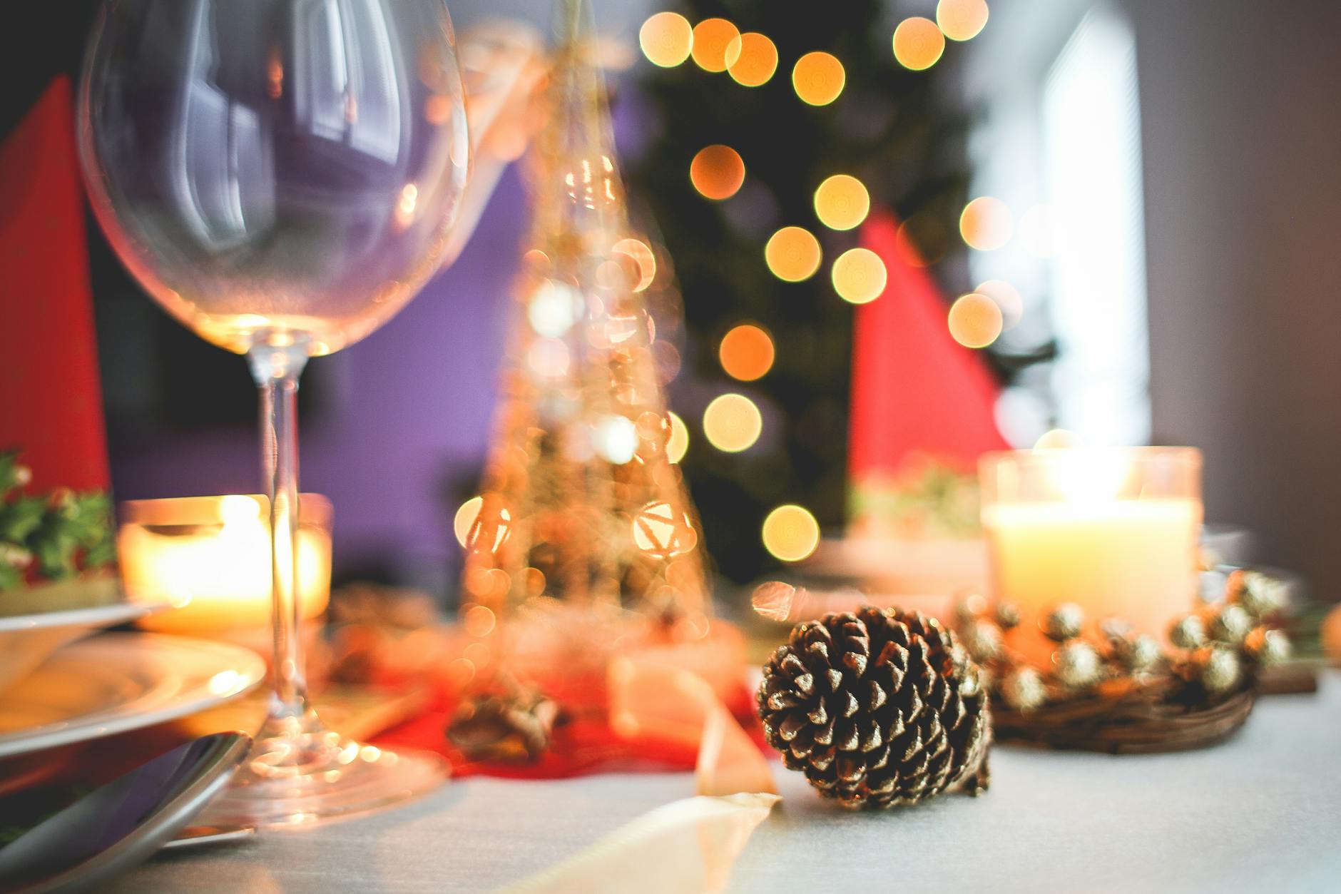 A candle sits on a softly styled table beside a pine cone, with a blurred Christmas tree glowing in the background. The scene evokes seasonal transition, warmth, and quiet anticipation.
