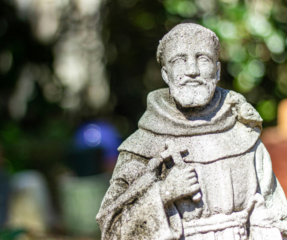 A serene statue of Saint Vincent the Martyr, depicted as a young deacon in a white tunic and red dalmatic, holding a palm branch and a book. His expression is peaceful, eyes lifted toward heaven. A faint halo glows around his head. In the background, soft light filters through a stone archway, suggesting both confinement and divine presence.